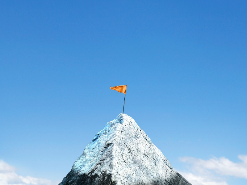 An orange flag is planted on top of a snow cap peak. Soft clouds frame the lower portion of the image and a rich blue sky.