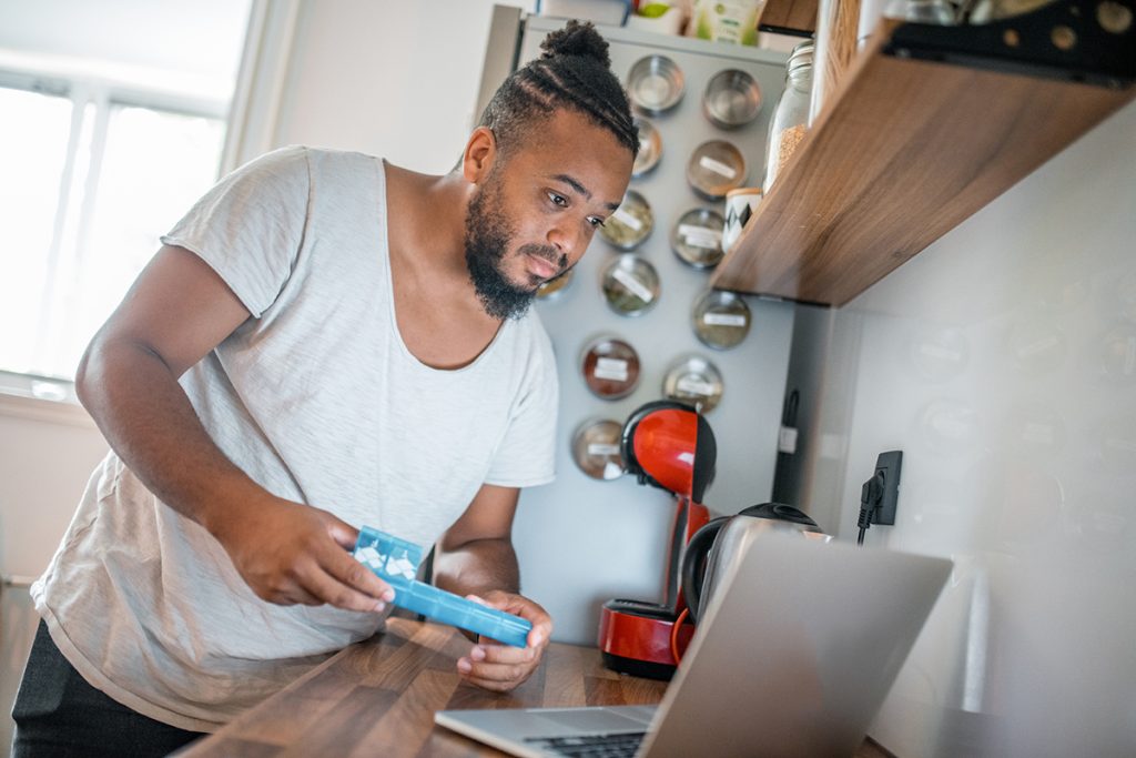 Man using laptop in the kitchen and holding a pill box in his hands