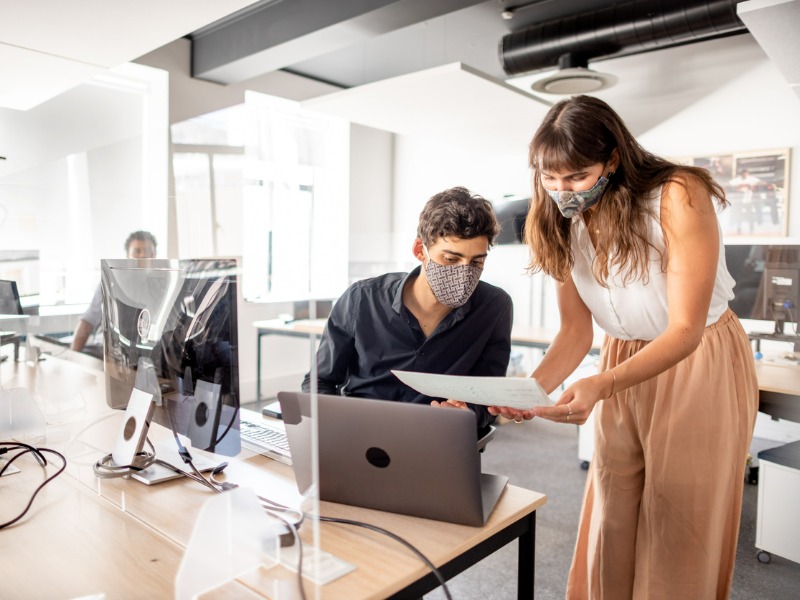 Two young businesspeople wearing protective face mask discussing paperwork together at a desk in a modern office