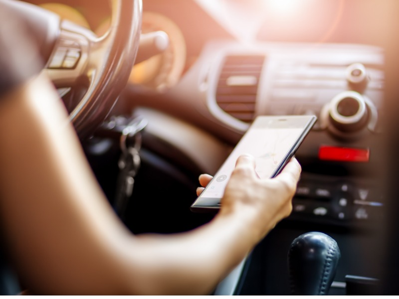 Driver driving a car on asphalt road in summer day at park. Woman at steering wheel with smartphone in her hand.