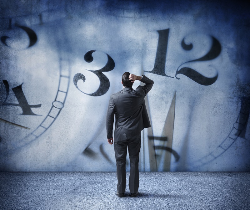 A rear view of a businessman as he places his hand on his head and looks up at random numbers from a clock face that appear on the wall in front of him.