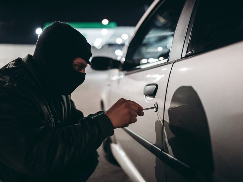 Man with a ski mask inserts a tool into a locked car door at night