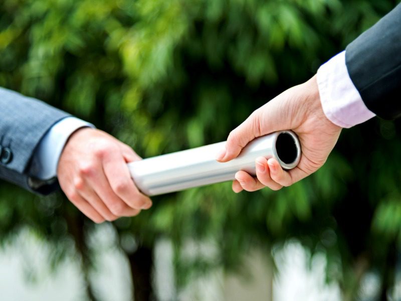 One businessperson in a grey suit is pictured handing a silver baton over to another businessperson in a black suit.