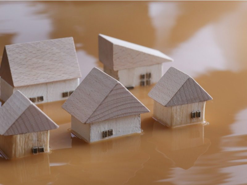 Five small wooden toy block houses are surrounded by murky brown water, up to their windows.