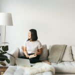 Smiling Asian Female Sitting On Sofa Working From Home In Minimalist Space
