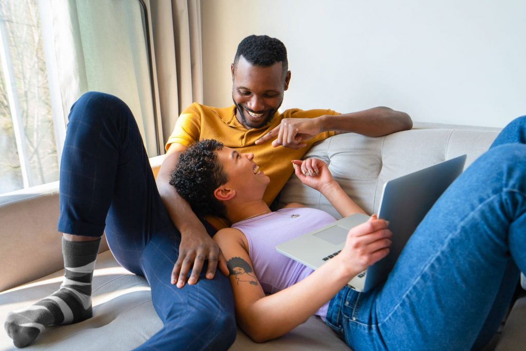 Black couple laughing on the couch with the woman holding a laptop and doing research