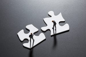 Two businessmen figurines each standing atop a white puzzle piece and looking pensive