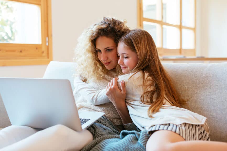 happy attractive young adult woman sitting on couch with her daughter with laptop on lap, smiling