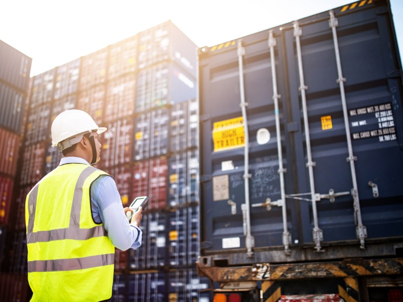 man wearing a white hardhat and yellow safety vest is onlooking a large blue shipping container on a truck