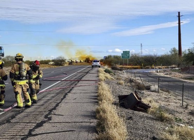 Firefighters stand on a highway as a orange smoke rises in the distance.