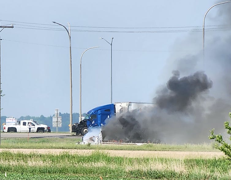 smoke rises from the scene of a road accident. a damaged blue semi-truck is seen in the background.