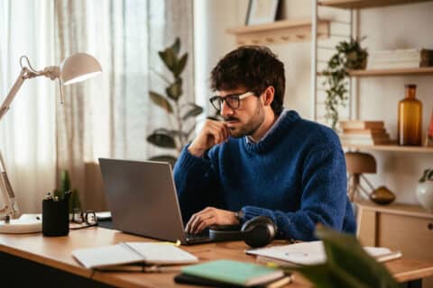 Smart bearded male freelancer in sweater and glasses touching face and thinking over data while sitting at desk and using laptop in light workplace at home.