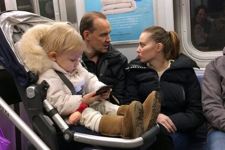 A baby in a pram stares at a phone on a subway carriage.