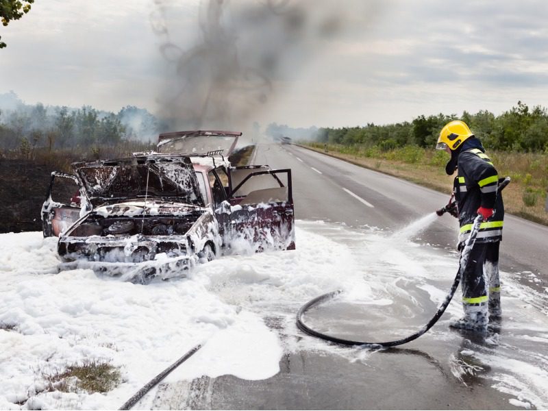 A firefighter using firefighting foam to spray a wrecked car on the side of the road