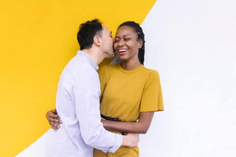 Tender boyfriend kissing smiling black girlfriend while standing on background of two colored wall in city