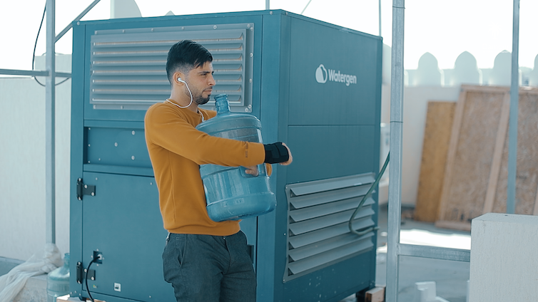 A mad carries a large water container past a cube-shaped machine.