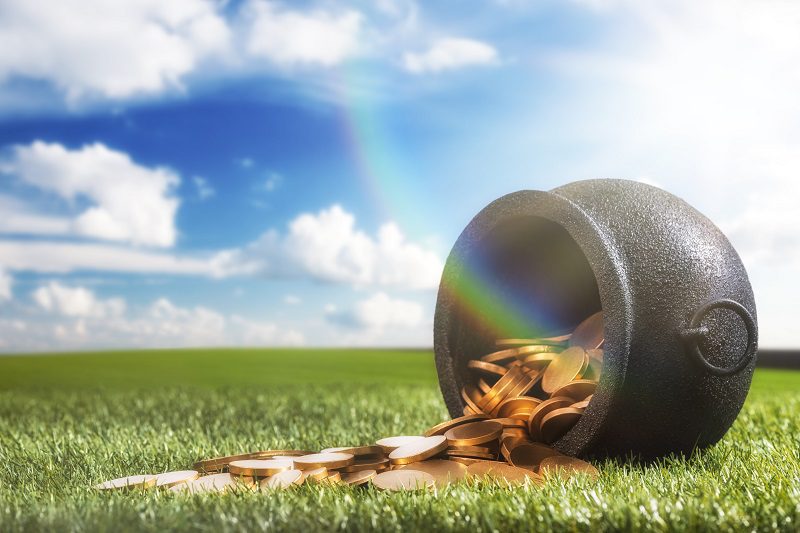 Low angle of a black wrought iron kettle pot that has been knocked over spilling it's gold coins onto the grass, with sun flare and rainbow. Mythology says it is the secret hiding place of an Irish Leprechaun's pot of gold, some even say he is tricky.