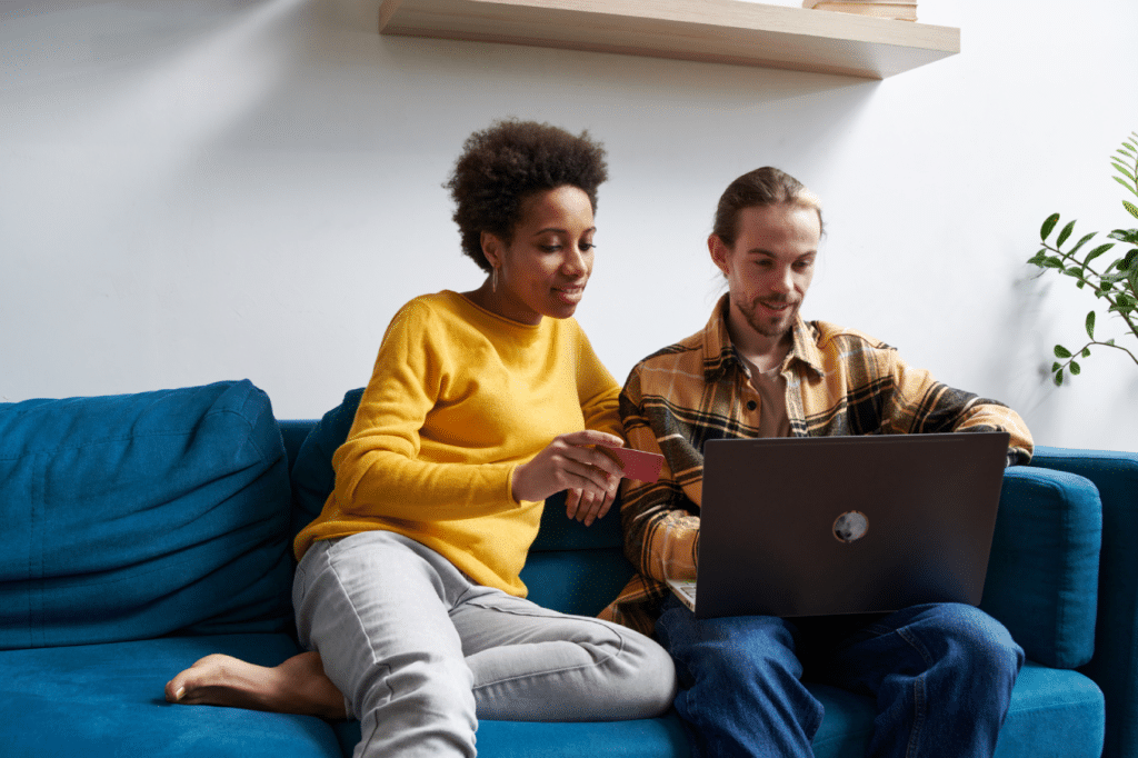 Contemporary couple relaxing at home and making online payment with plastic card while shopping on netbook