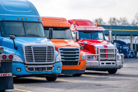 Different bonnet makes and models of professional big rigs semi trucks with commercial cargo on semi trailers standing in row on the industrial truck stop parking lot waiting for unloading