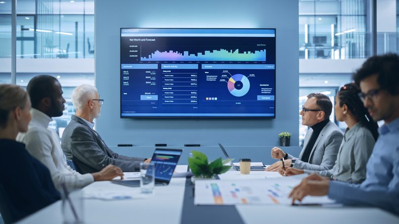 Six business leaders sit at a conference table looking toward a software presentation on screen