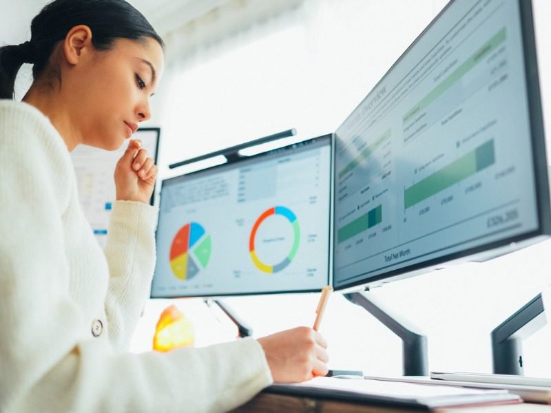 Woman working with two desktop screens that are displaying circle and bar graph data