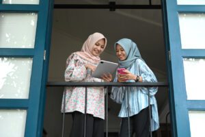 A woman in a pink hijab smiles and points to the tablet in her hand, while a woman in a blue hijab looks over her shoulder