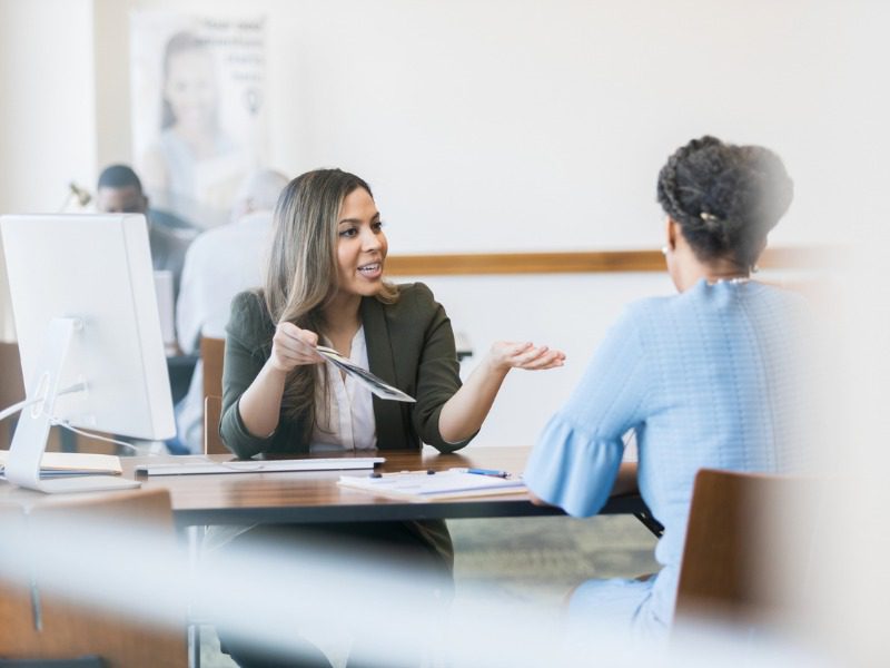 Female insurance professional sits across a desk and gestures in discussion with a female customer.