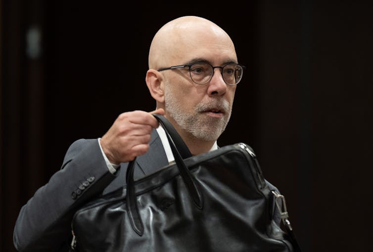 A bald white man wearing glasses and a grey suit carries a black briefcase