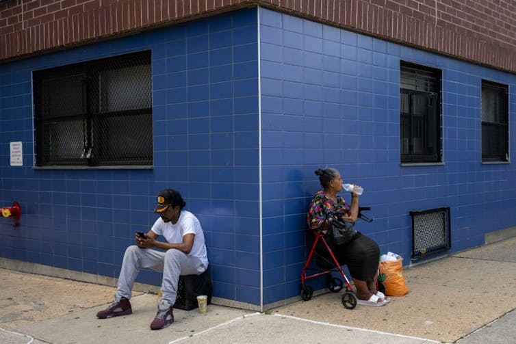 Two people sitting with their backs against the corner of a blue building,