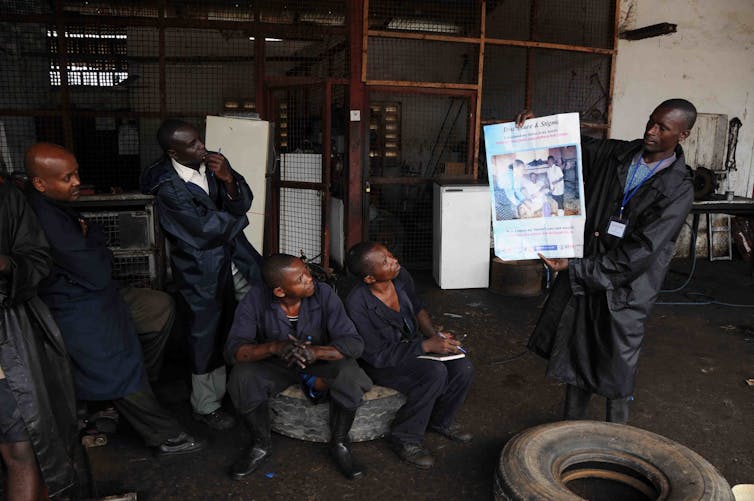 several men wearing coveralls watch another man who's holding up a poster